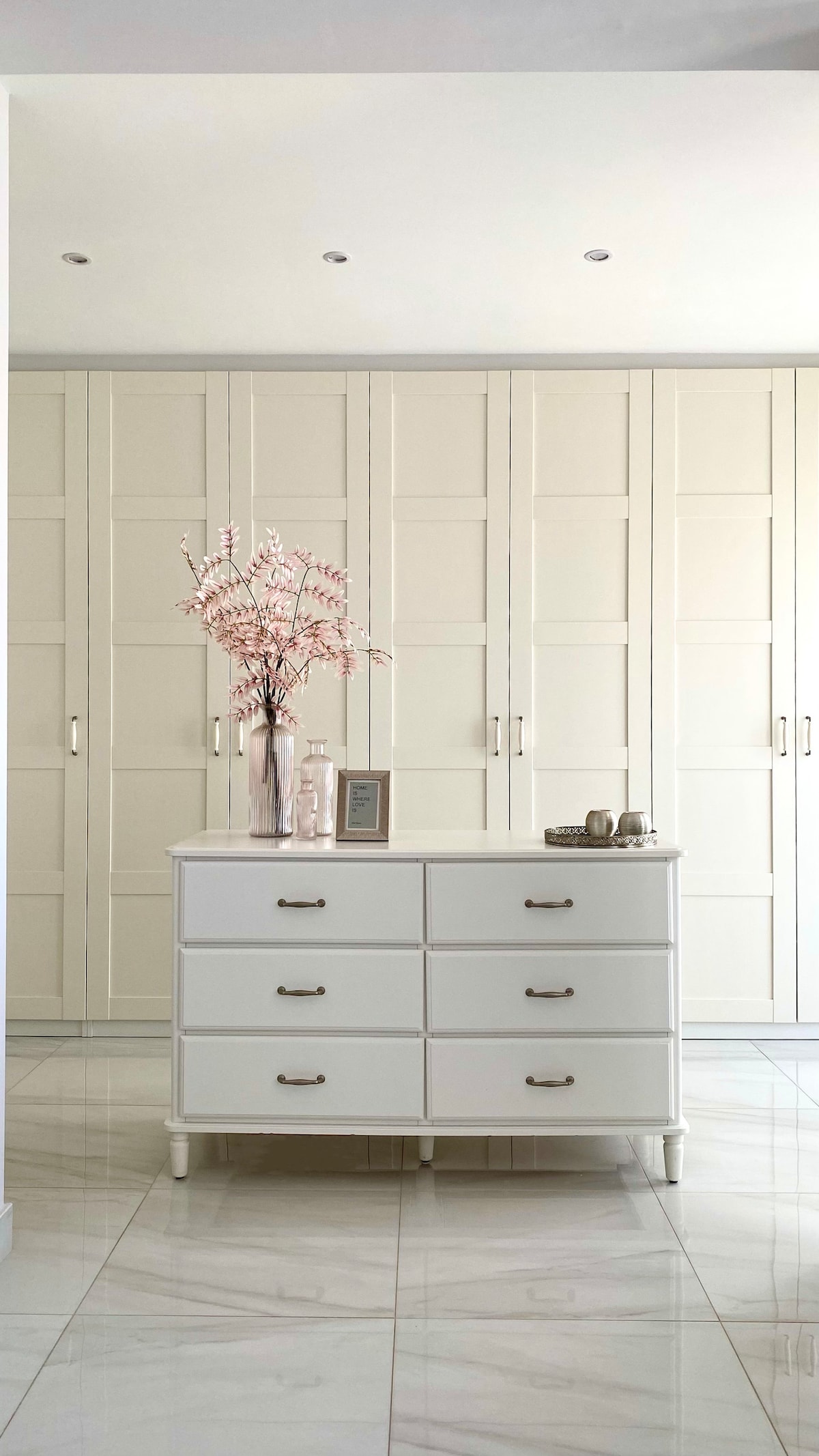 A bright entryway is showcased, featuring a white dresser with six drawers. A minimalist vase with delicate pink branches sits on top. Behind, sleek white cabinetry complements the clean aesthetic, with recessed lighting above illuminating the space subtly.