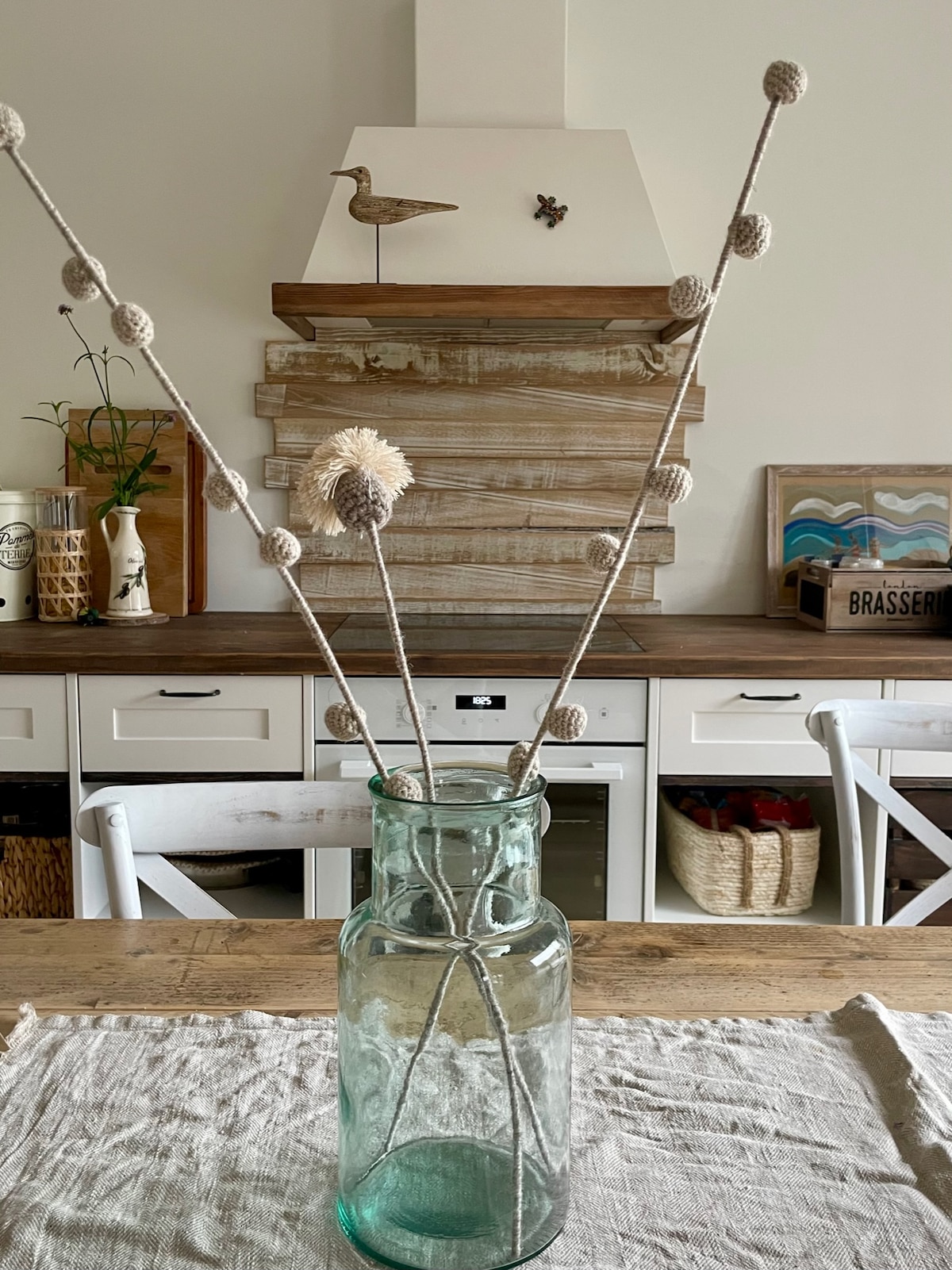 A glass vase filled with decorative branches stands on a wooden table. The branches feature round plant accents and are artistically arranged. A neutral-toned tablecloth covers the surface, with a wooden kitchen backdrop visible, enhancing a simple and natural aesthetic.