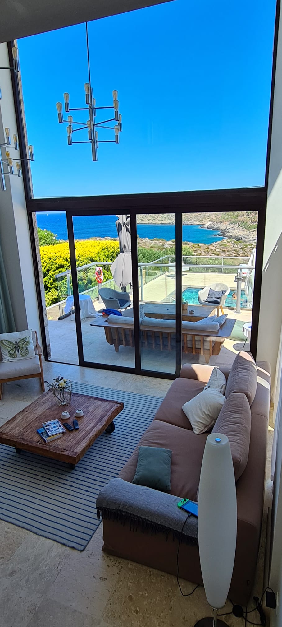 A spacious living area is showcased with large glass doors that open to stunning sea views. A light brown sofa sits elegantly on a textured rug, accompanied by a wooden coffee table. A modern light fixture hangs from the high ceiling, enhancing the airy atmosphere.