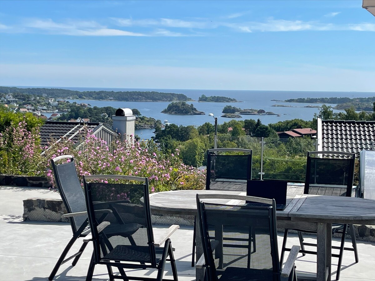 An outdoor terrace features a large wooden table surrounded by several sleek black chairs. A panoramic view of the sea and nearby islands is visible, with lush greenery and flowering plants framing the scene under a clear blue sky.
