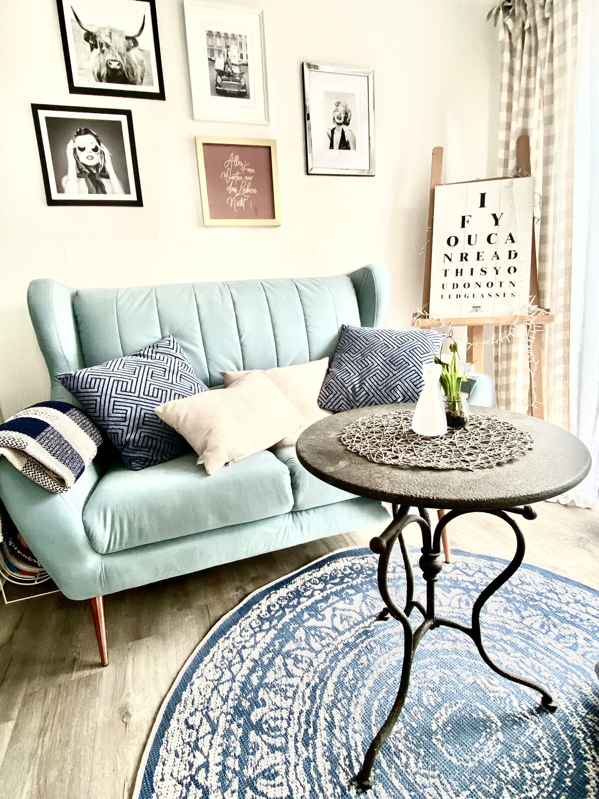 A cozy seating area features a light blue sofa adorned with patterned cushions. A round table stands beside it, topped with a small vase holding fresh flowers. A decorative rug adds texture to the space, complemented by framed artwork on the walls.