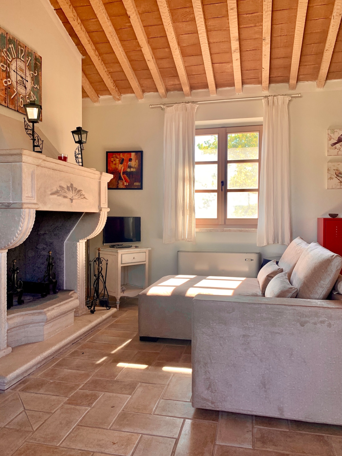 A spacious living area is highlighted by a stone fireplace and a plush sofa. Natural light filters through a large window adorned with white curtains, illuminating the warm, earthy tones of the tiled floor. Decorative artwork adds a personal touch to the space.