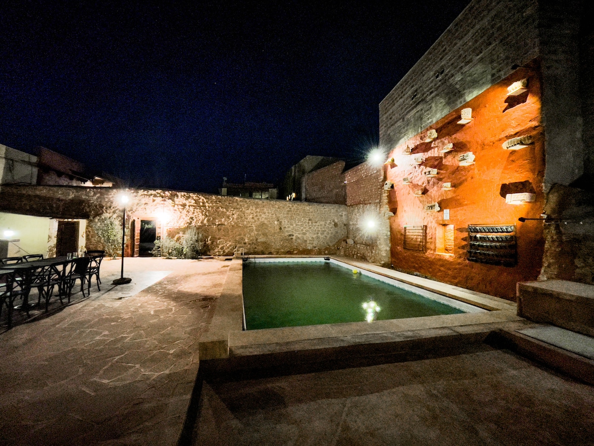 A serene outdoor area features a rectangular pool surrounded by stone pathways. The wall beside the pool has artistic elements and is softly lit by ground-level lights, enhancing the evening ambiance. A dining table with multiple chairs stands nearby, ready for gatherings.