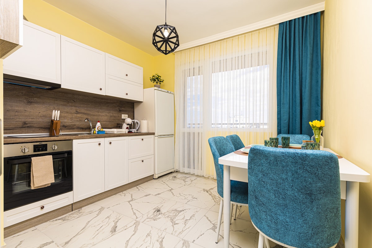 The kitchen features modern white cabinetry against a yellow wall, with a tiled floor in a light pattern. A dining table with four teal chairs is set near a window dressed with sheer white curtains. Appliances include an oven, fridge, and kettle.