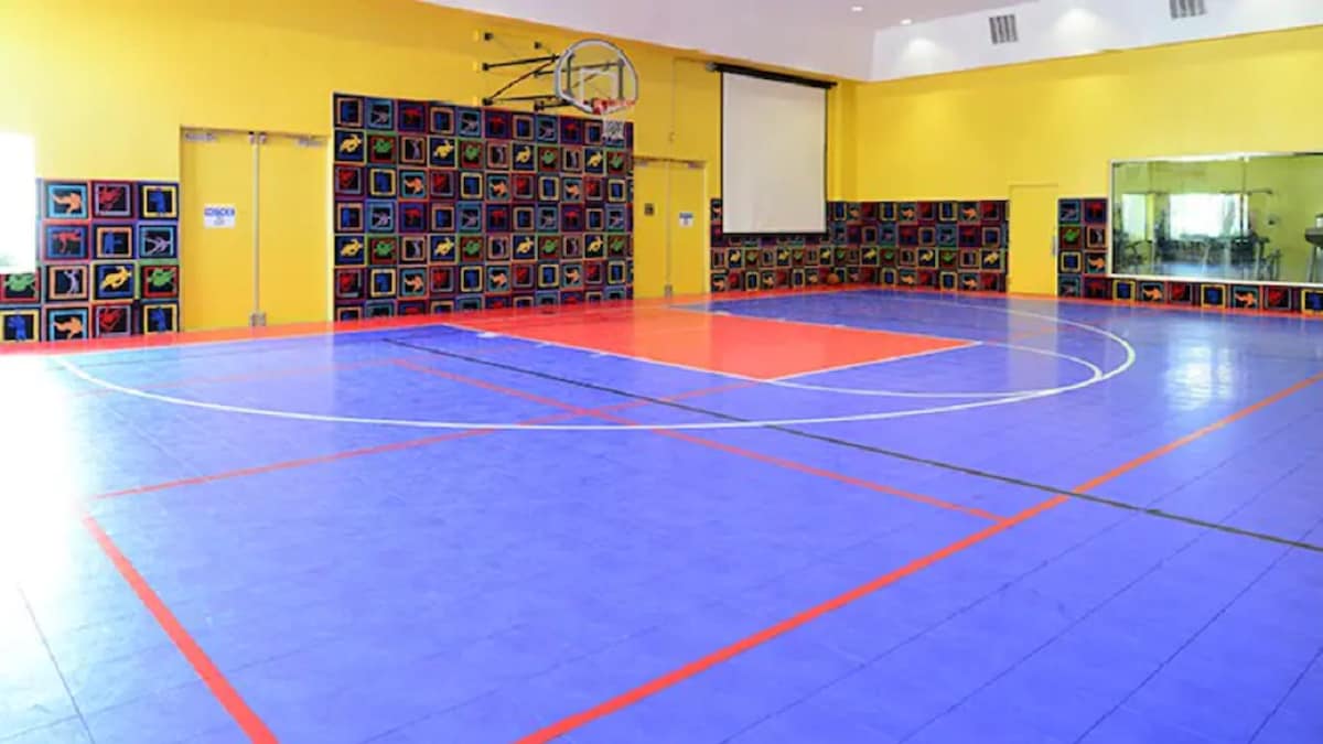 A vibrant indoor sports area features a brightly colored court with a blue surface and marked play zones. Surrounding walls are adorned with colorful artwork, and a basketball hoop is mounted overhead. A large mirrored wall reflects the space, enhancing its openness.