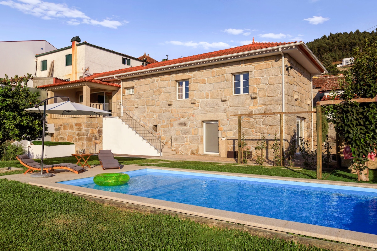 A traditional stone house is set against a clear sky, featuring a private swimming pool with a vibrant green float. Lush grass surrounds the pool area, and a lounge chair provides a space for relaxation. The exterior is complemented by minimalist garden features.