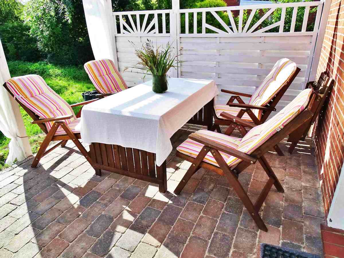 An outdoor seating area features a rectangular wooden table surrounded by four lounge chairs with striped cushions. A vase with greenery rests on the table, and natural light filters through the outdoor space, creating a relaxed setting for gatherings.