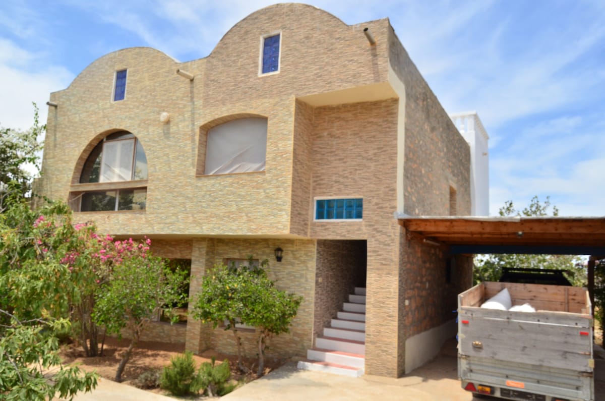 The exterior of the guest house displays a unique architectural design with arched windows and a textured stone facade. A stairway leads to the entrance, surrounded by greenery and flowers. A covered area for parking is visible on the right, offering convenience for guests.
