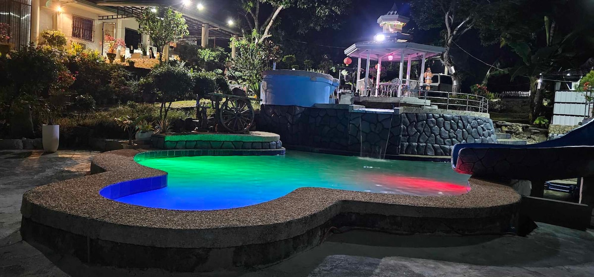 A spacious pool is illuminated with colorful lights, creating a vibrant atmosphere at night. Surrounding greenery complements the scene, while a covered seating area is visible in the background. The pool's unique shape adds charm, encouraging relaxation and enjoyment.