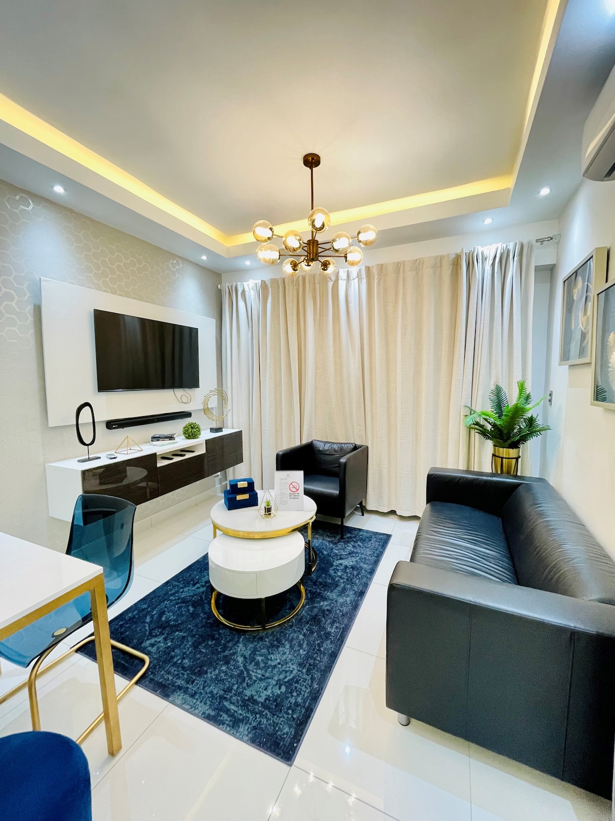 A modern living space features a black leather sofa paired with a sleek armchair. A round coffee table sits on a navy area rug, with a decorative plant positioned beside it. Soft lighting is provided by a stylish chandelier, complemented by draped curtains and a wall-mounted television.