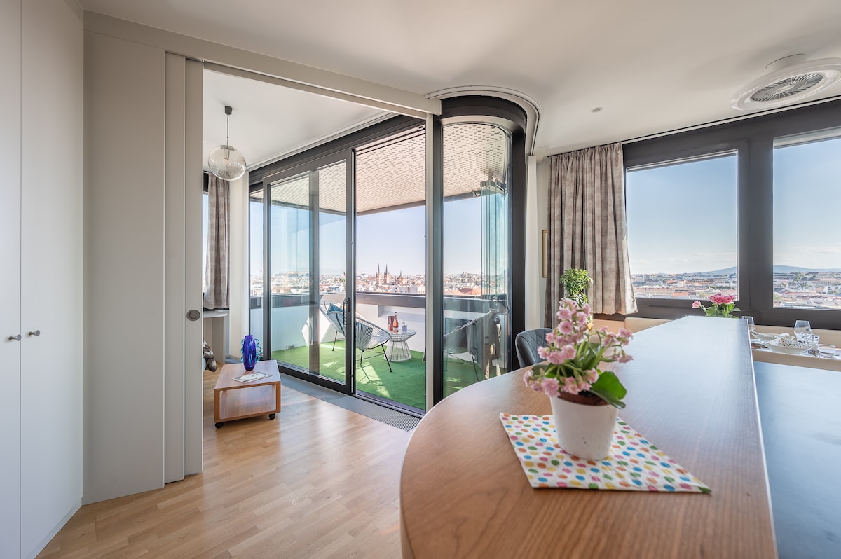 An open living space connects to a spacious loggia, featuring floor-to-ceiling glass doors that provide a panoramic view of the city. A small potted plant decorates the countertop, while comfortable outdoor lounge chairs are set against a backdrop of vibrant greenery on the balcony.