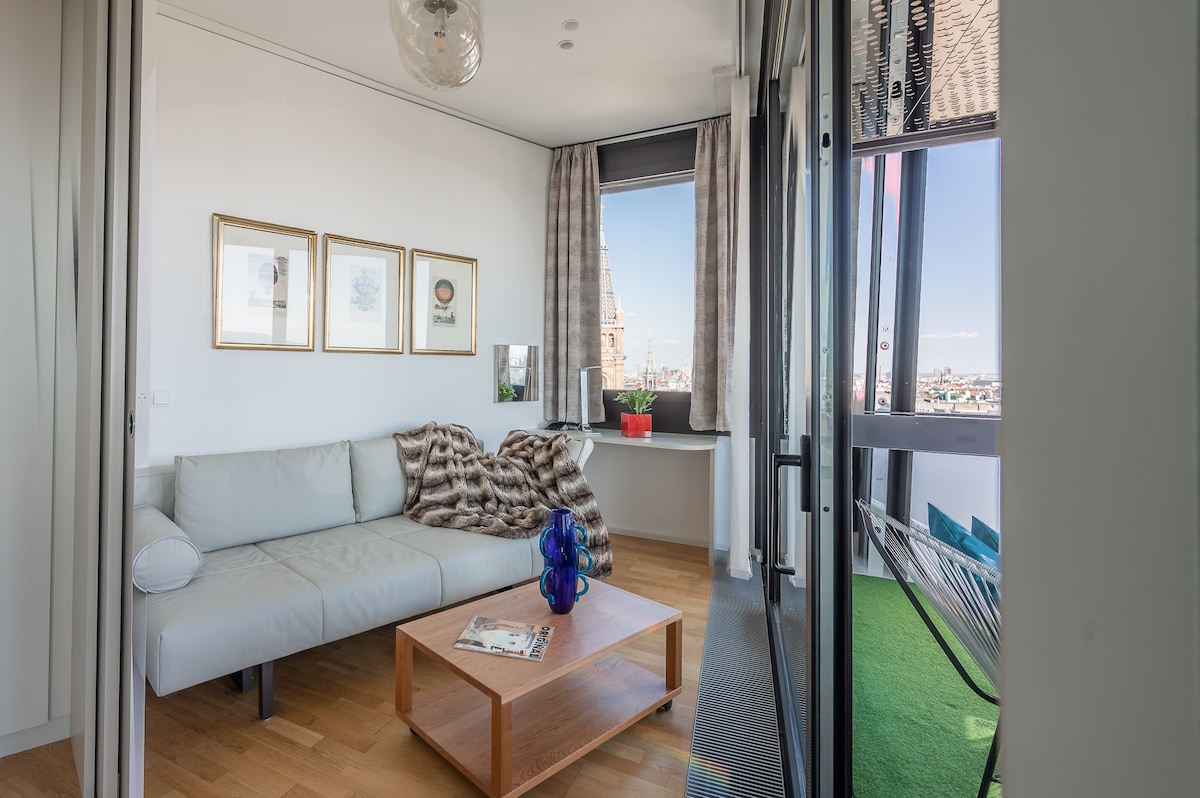 A spacious living area features a light-colored sectional sofa paired with a wooden coffee table. Nearby, a large window showcases city views, while soft curtains frame the scene. Decorative artwork is displayed on the wall, adding a touch of style to the room.