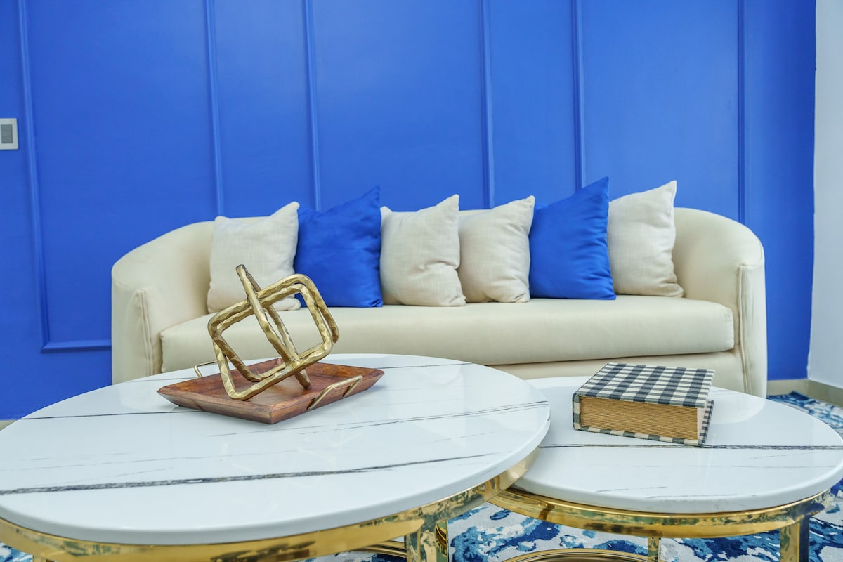 A cozy seating area features a light-colored sofa adorned with cream and blue pillows. Two round coffee tables with marble tops are positioned in front, accompanied by a decorative object and a book, set against a vibrant blue wall.