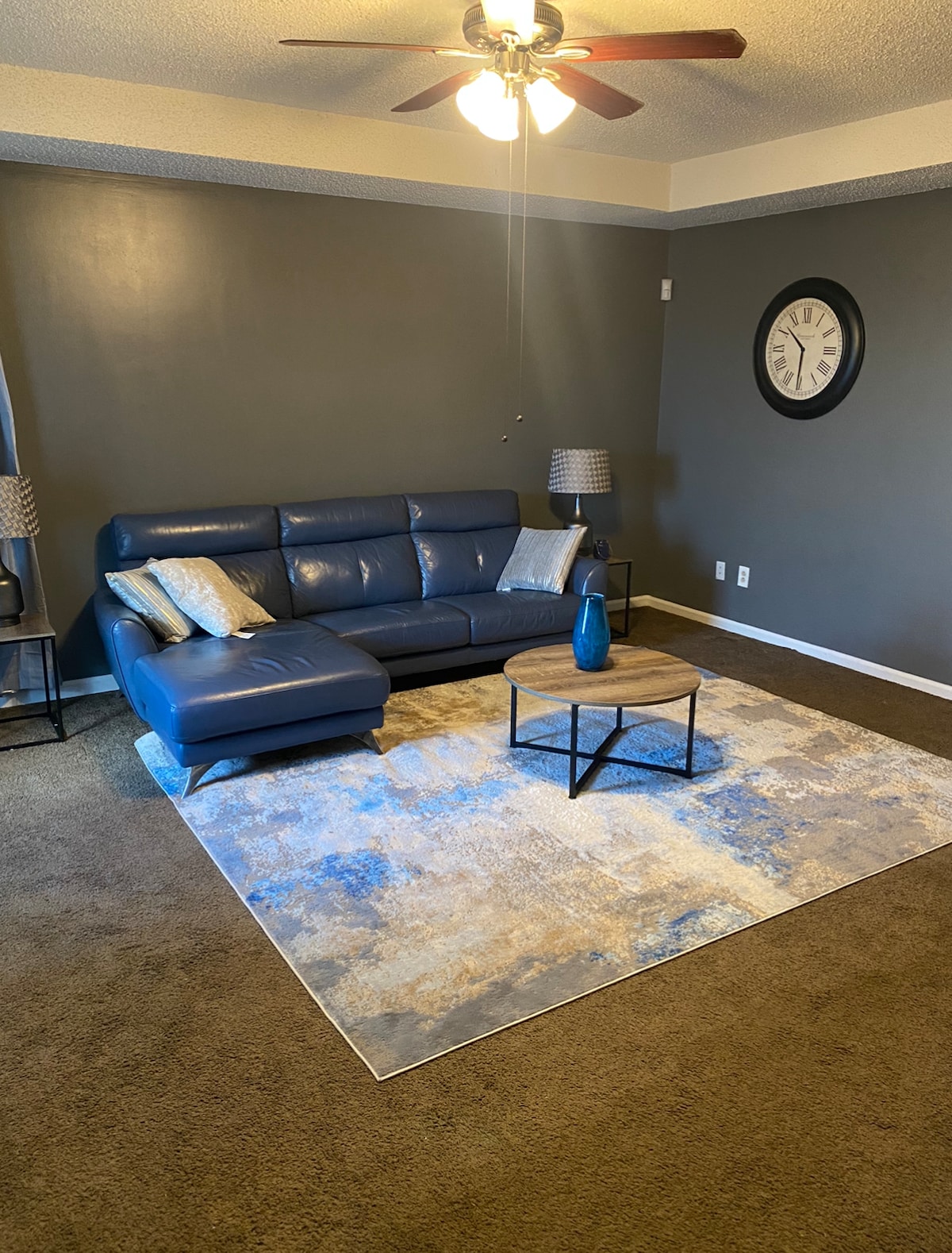 A living area features a blue sectional sofa paired with two decorative lamps on either side. A round coffee table sits on a patterned area rug that complements the room's subtle color scheme. A wall clock adds a functional touch to the space.