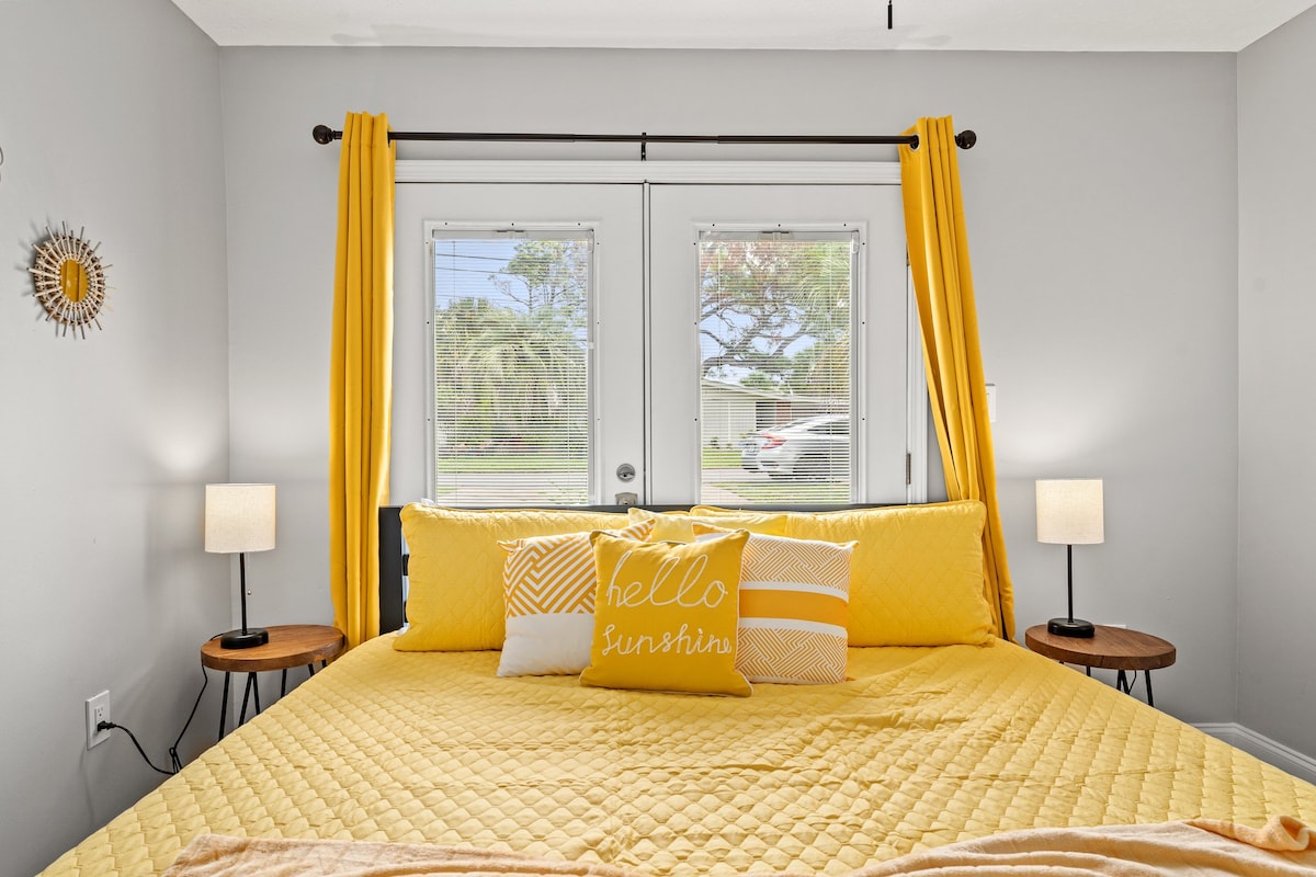A bright and welcoming bedroom features a spacious yellow bedspread with matching decorative pillows. Natural light enters through double doors adorned with yellow curtains, and bedside tables hold soft-lit lamps. A cheerful wall decoration adds a touch of warmth to the room's neutral tones.
