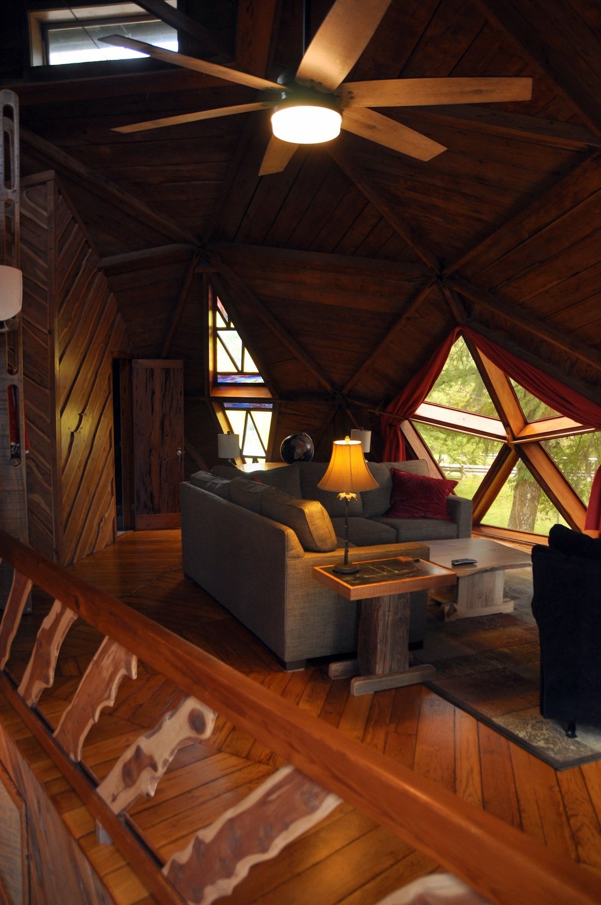 A cozy living area is highlighted by a unique geodesic design, featuring a large window with triangular panes. A sofa and armchair are arranged around a natural wood coffee table, with warm light from a lamp creating an inviting atmosphere.