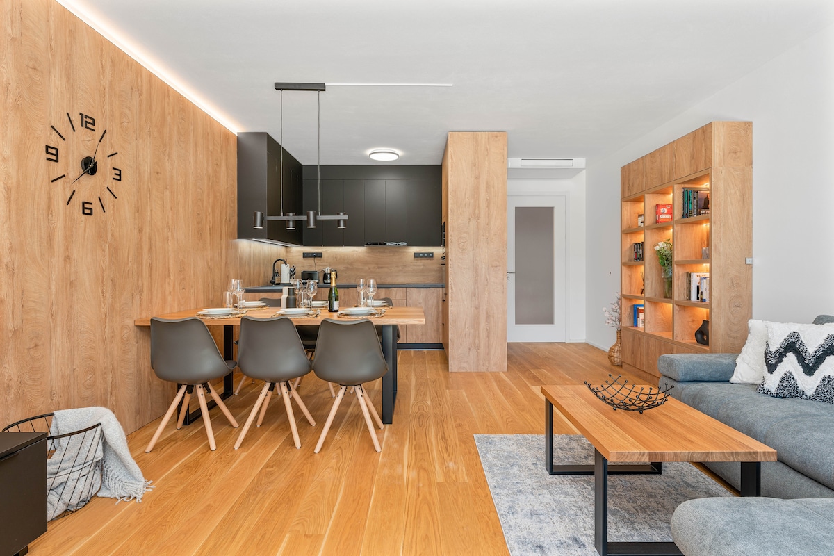 An open-concept kitchen and dining area features a modern wooden dining table set for four. The kitchen is equipped with dark cabinetry and stainless steel appliances. A comfortable seating area includes a deep gray sofa and a coffee table. Warm wood accents enhance the space.