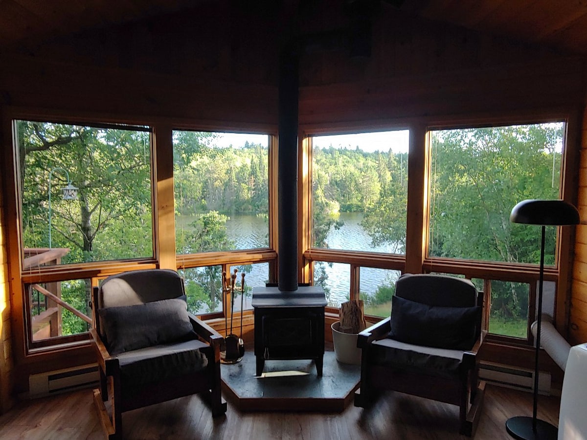 A cozy sitting area is framed by large windows that overlook the lake and surrounding greenery. Two comfortable armchairs are positioned near a wood-burning stove, creating a peaceful retreat, while natural light fills the space, enhancing the connection with nature outside.