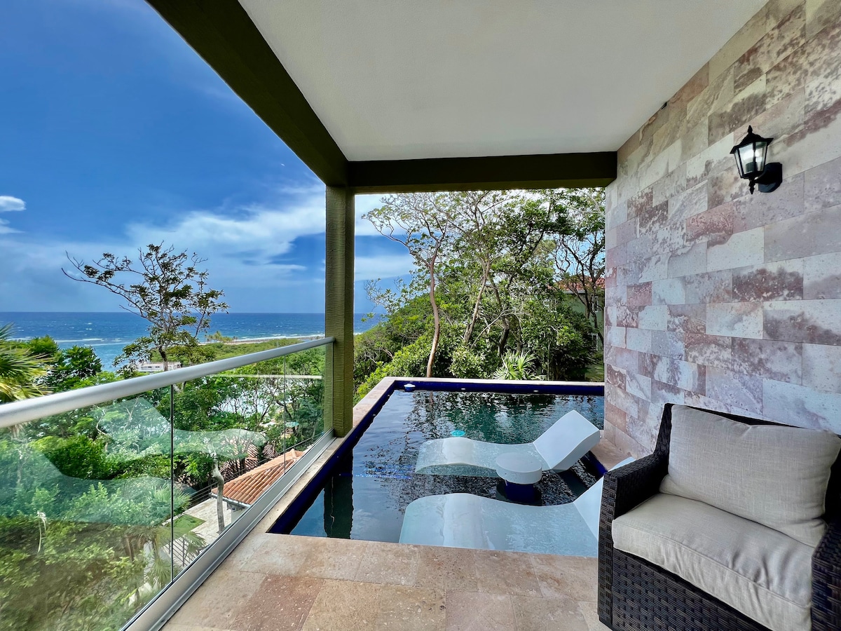 An inviting private pool is seen on a covered terrace, featuring two lounging chairs positioned for relaxation. Glass railings provide unobstructed views of the surrounding greenery and the ocean beyond, with a bright blue sky overhead. Natural light fills the space, enhancing the serene atmosphere.