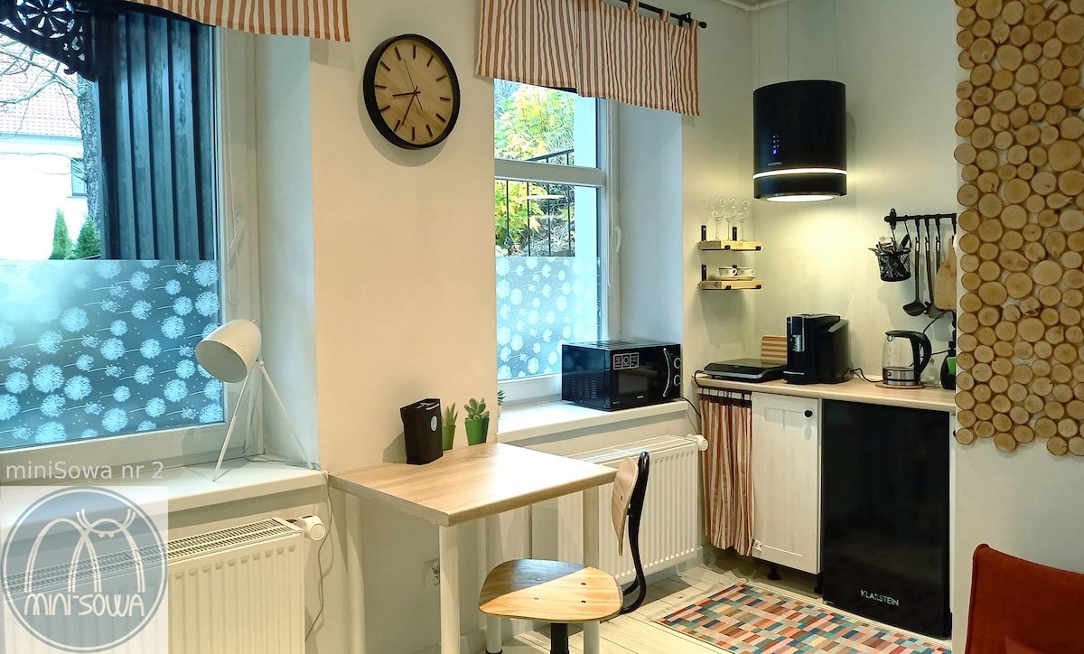A compact kitchen area features a microwave and coffee maker against a backdrop of wooden accents. A small table with a chair is positioned near the window, which is adorned with light curtains. A wall clock and stylish lighting add to the functional design.