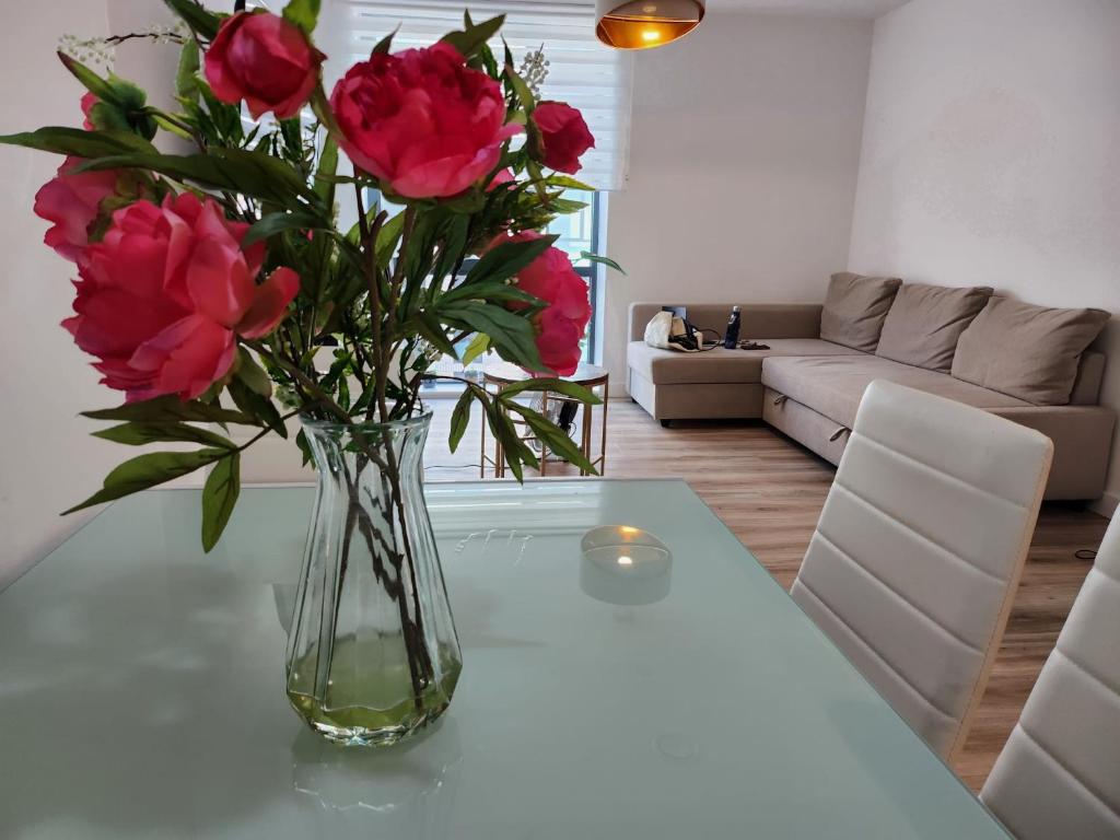 A glass dining table is set with a clear vase filled with vibrant pink flowers. In the background, a neutral-toned sofa is visible, accompanied by a side table that includes a drink. Soft lighting illuminates the inviting space.