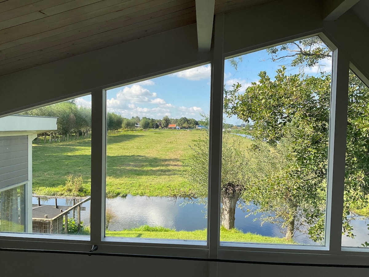 Large windows provide a panoramic view of the surrounding farmland and water, allowing natural light to fill the space. The landscape features lush greenery and a serene body of water, creating a tranquil atmosphere. The sloped roof is visible in the foreground.