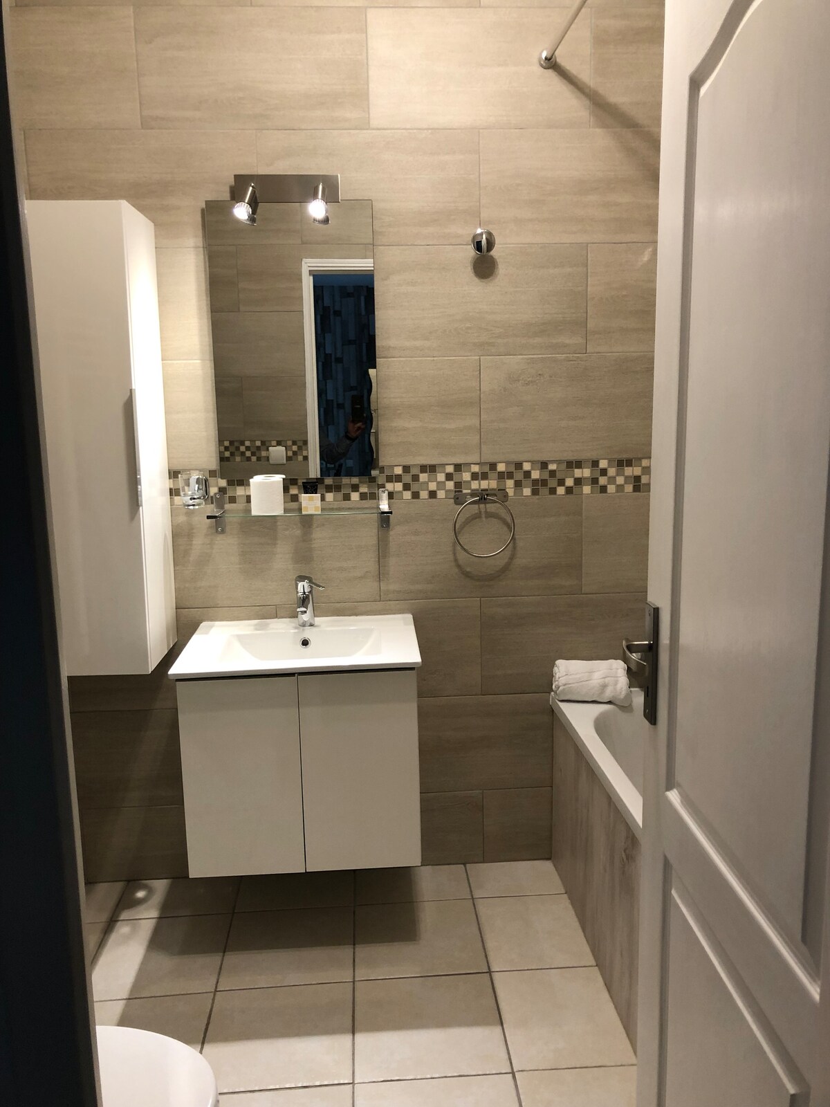 The bathroom features modern tilework in neutral tones and a large mirror above a sleek vanity with storage. A bathtub with a shower fixture is positioned alongside the wall. Towels are neatly arranged on a bar, and a wall-mounted holder is visible.
