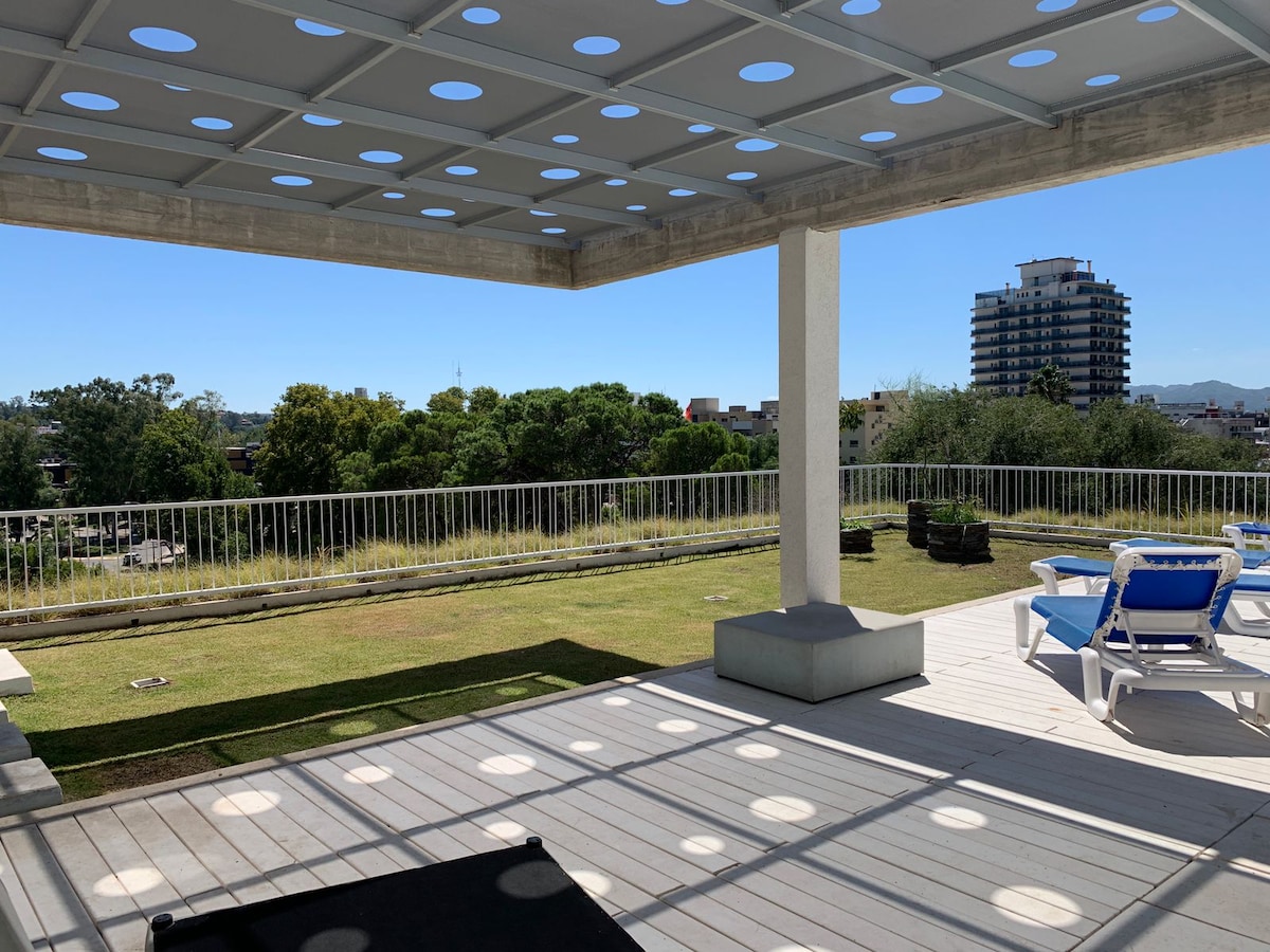 An outdoor terrace features a shaded area with a patterned roof and comfortable loungers. A view of the lush greenery and nearby buildings is visible in the background, while a manicured lawn extends across the space.