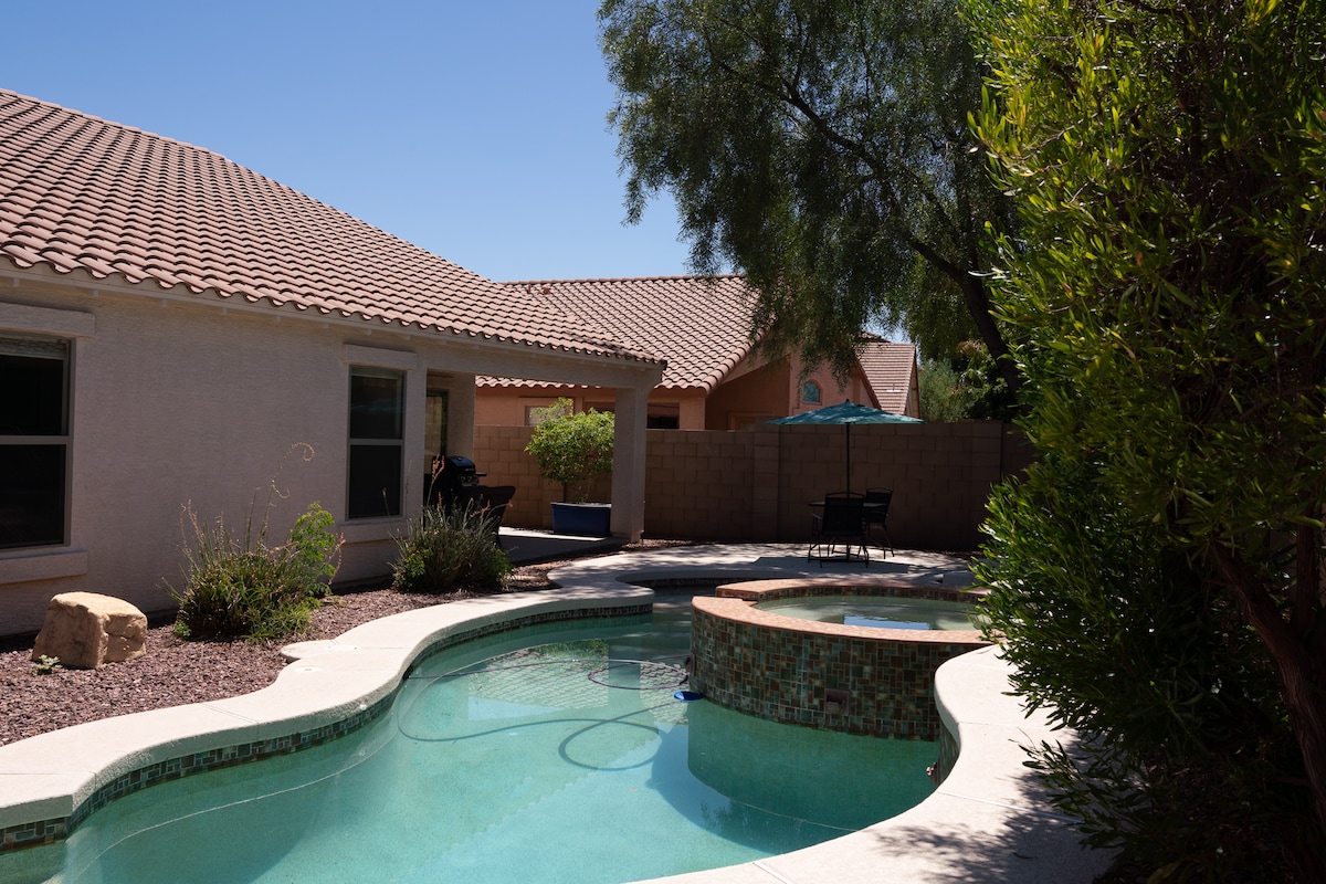 A tranquil outdoor space features a curved pool surrounded by lush greenery and smooth stone accents. A shaded area with an umbrella is visible in the background, offering a spot for relaxation. The warm sunlight reflects off the water's surface, enhancing the inviting atmosphere.