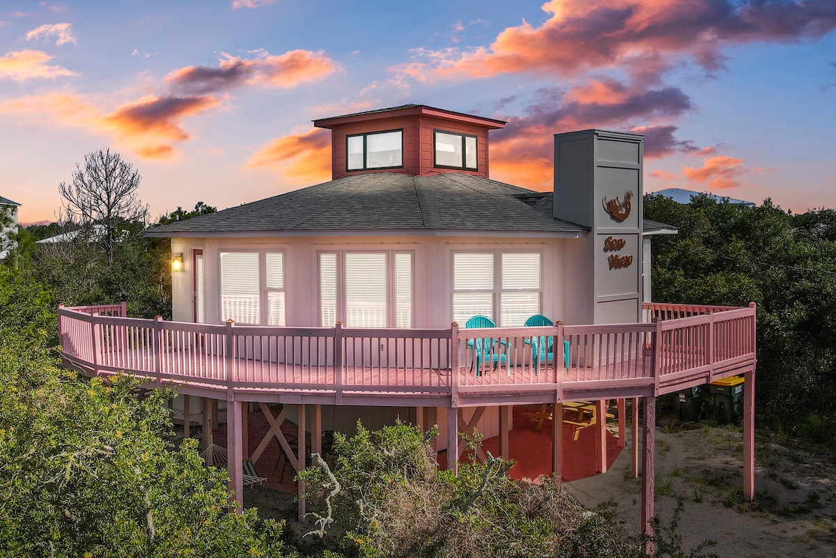 The exterior of the Sea View is presented, featuring a circular wraparound deck and a lighthouse structure on the roof. Two turquoise chairs are positioned on the deck, surrounded by a natural landscape, with a sunset sky in the background creating a soft glow.