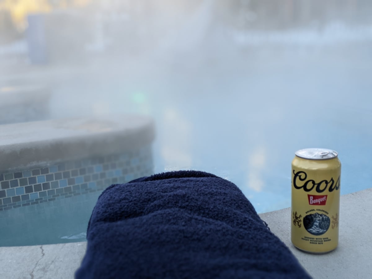 A towel is neatly placed beside a steaming outdoor hot tub, with soft mist rising around the area. A can of beer sits on the edge near the water, complementing the serene atmosphere.
