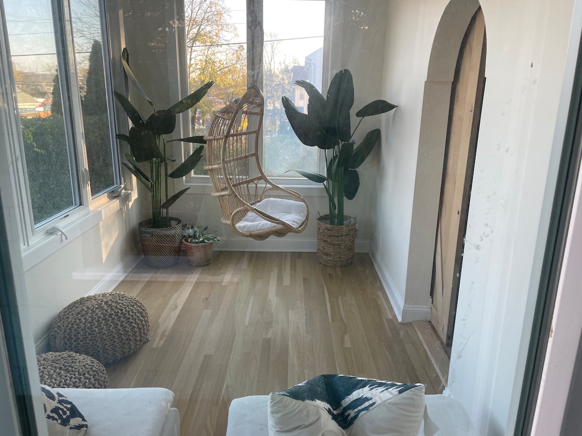 A bright sunroom features large windows allowing natural light to fill the space. A hanging chair is surrounded by leafy plants, complemented by textured poufs on the wooden floor. The room offers a serene atmosphere for relaxation or reading.