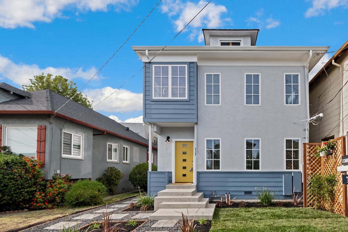 A modern two-story apartment building features a light gray exterior with distinctive blue accents. A bright yellow front door draws attention. Symmetrical windows provide ample natural light. A well-maintained pathway leads to the entrance, bordered by neatly arranged landscaping.