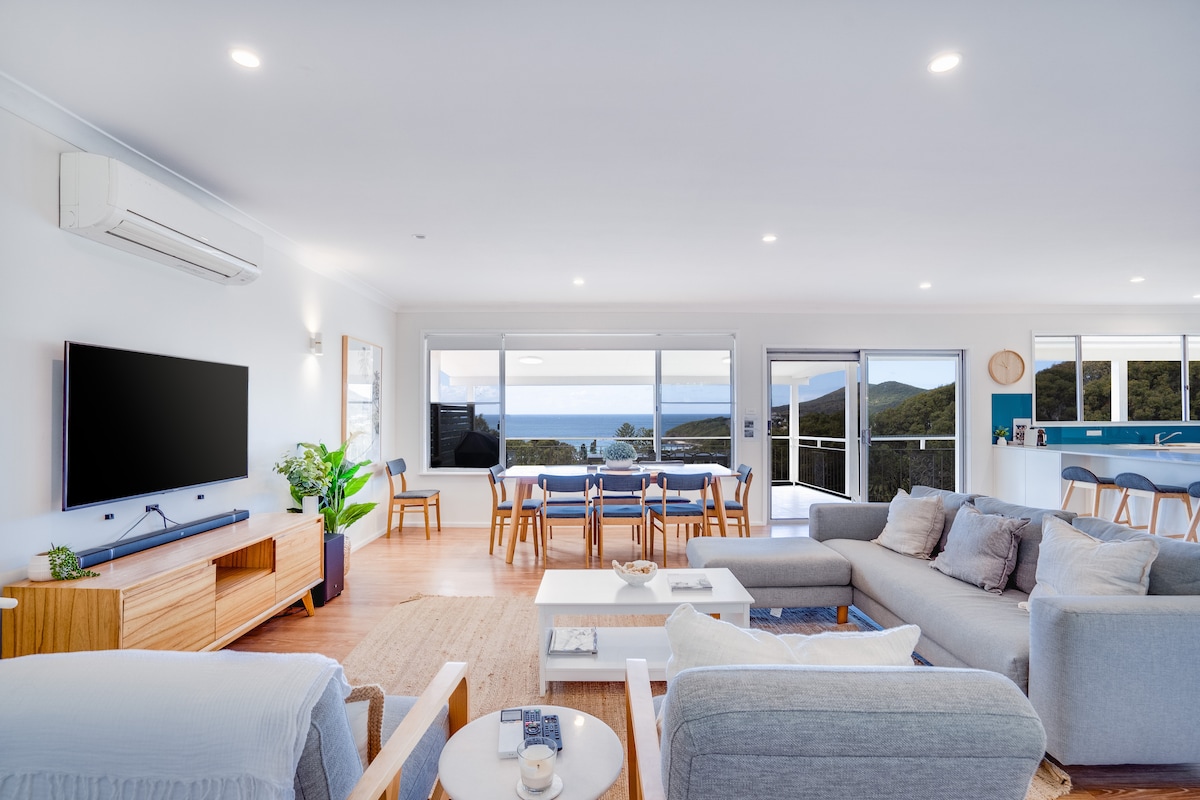 A spacious open plan living area features a large sofa and a minimalist coffee table. Natural light floods in through large windows, showcasing ocean views. A dining table for six is positioned nearby, and the kitchen is visible in the background.