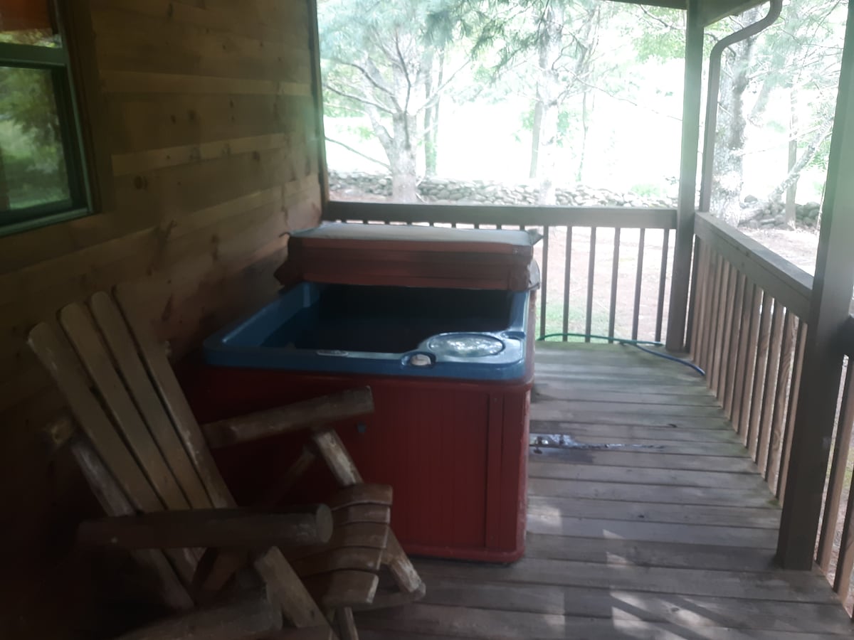 A private porch area features a Jacuzzi hot tub beside a wooden rocking chair. The surrounding space is framed by wooden railing and is bordered by trees, providing a peaceful setting for relaxation.