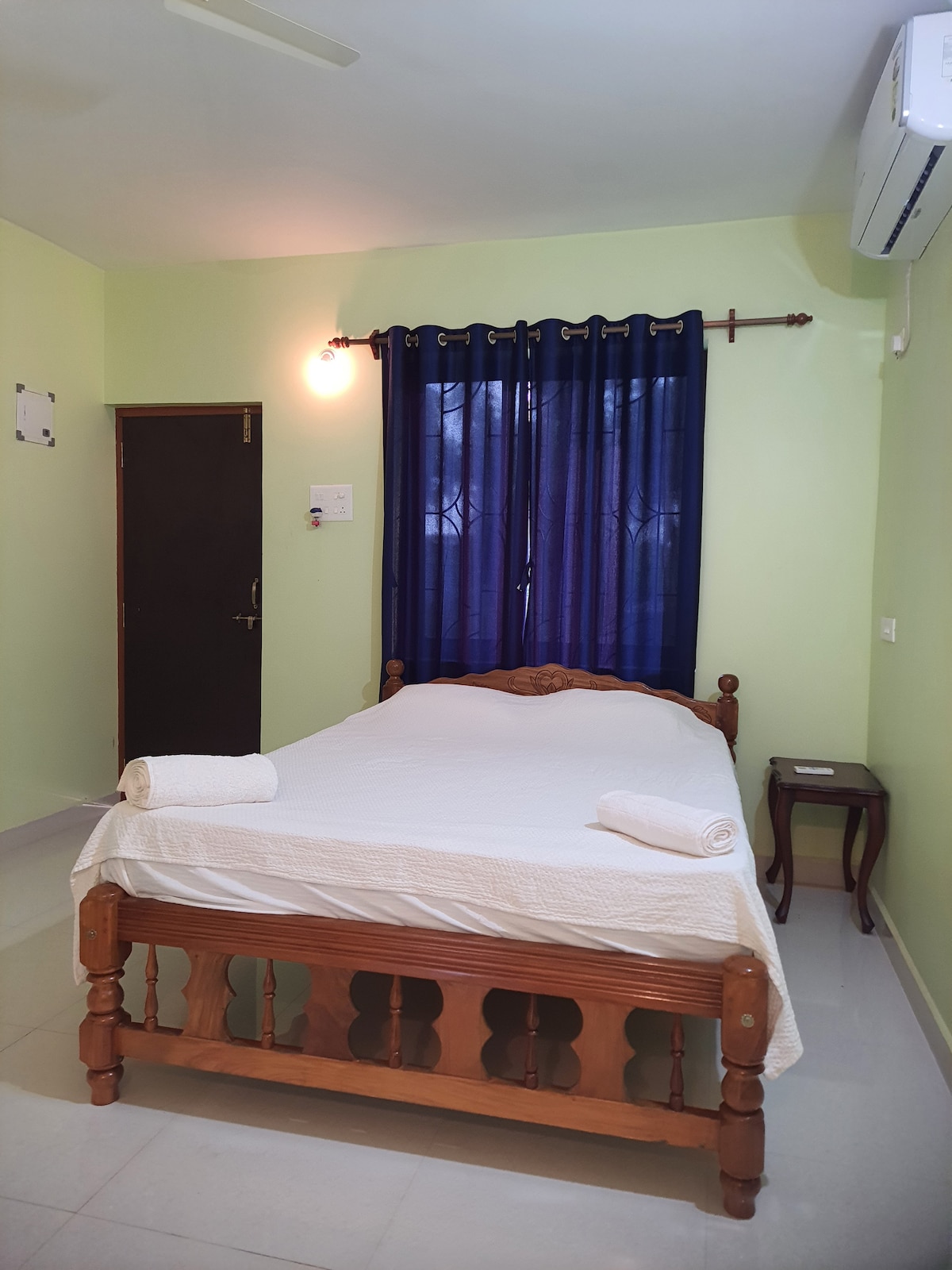 A well-lit bedroom features a wooden bed with a light-colored blanket. Two rolled towels are neatly placed on the bed. Dark curtains frame a window, and a small wooden nightstand is positioned beside the bed. A wall-mounted air conditioning unit is visible.