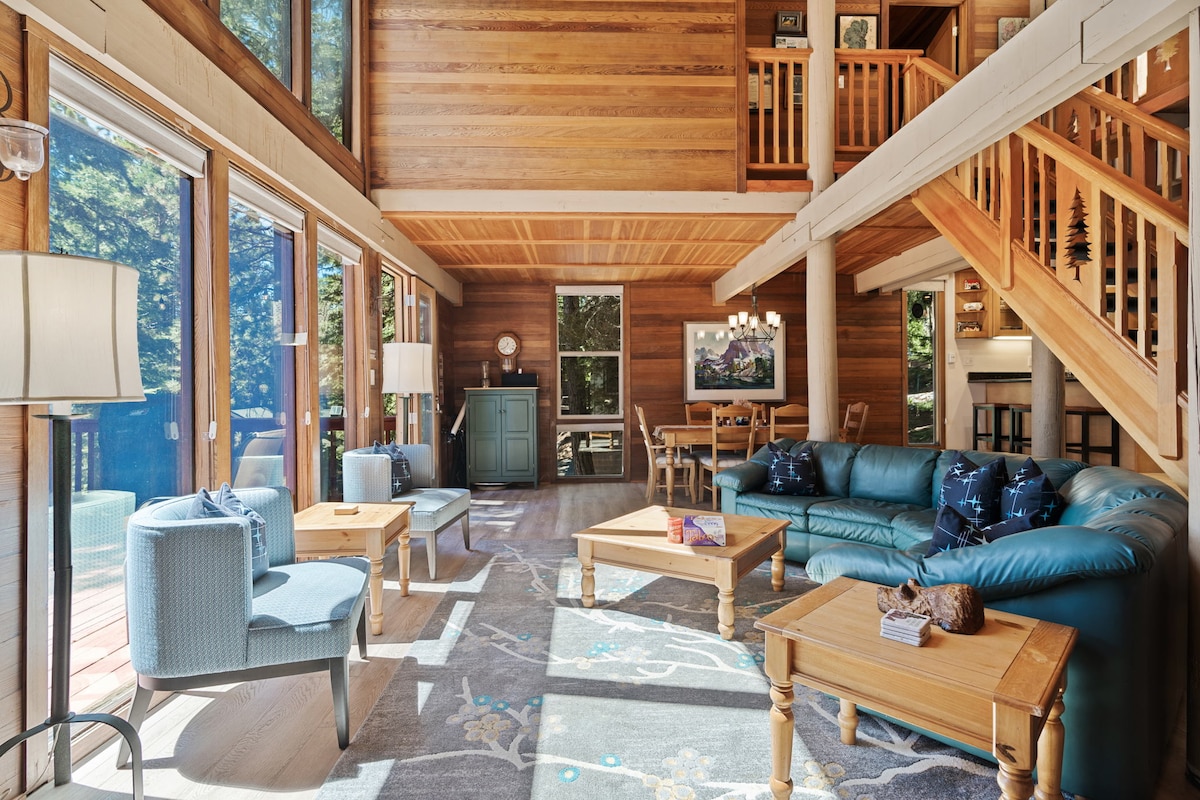 A spacious living area features high ceilings and large windows that provide a view of the surrounding pine trees. Natural light fills the room, highlighting comfortable seating arrangements and wooden furniture. A dining area is visible in the background, creating an inviting space for gatherings.
