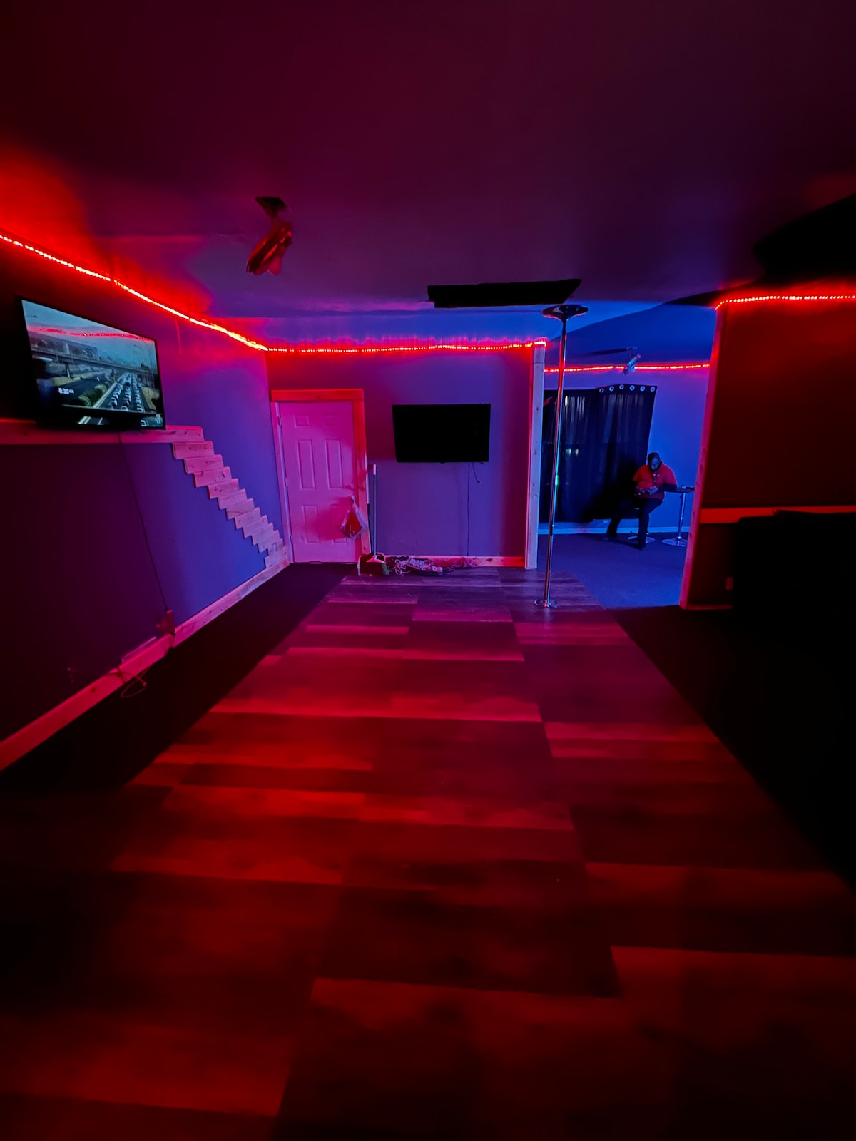 The interior area features a vibrant color scheme with red and blue LED lighting accentuating the walls. A pole stands in the center, and multiple screens are mounted on the walls. The floor is covered in wooden planks, contributing to the space's unique decor.