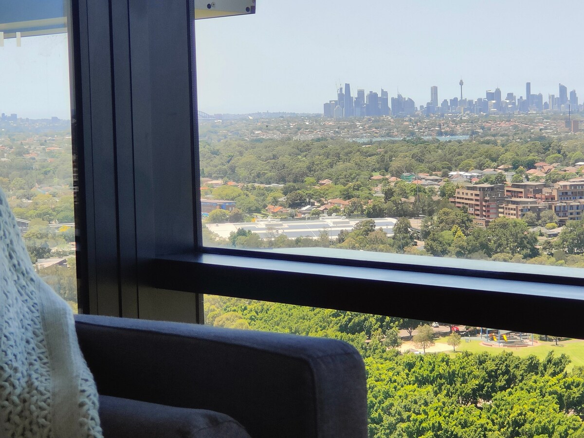 A clear view from a high floor showcases a sprawling urban landscape, merging patchy greenery with the distant city skyline. Large windows frame the scene, inviting natural light into the space. A cozy chair is positioned nearby, enhancing relaxation while enjoying the panoramic view.