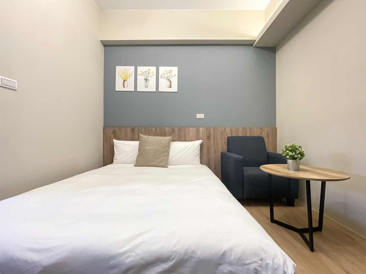 A clean and simple room features a spacious bed with white linens and a neutral-colored pillow. A small round table and a chair are placed nearby. Three framed floral prints adorn the blue accent wall, adding a touch of color to the neutral palette.