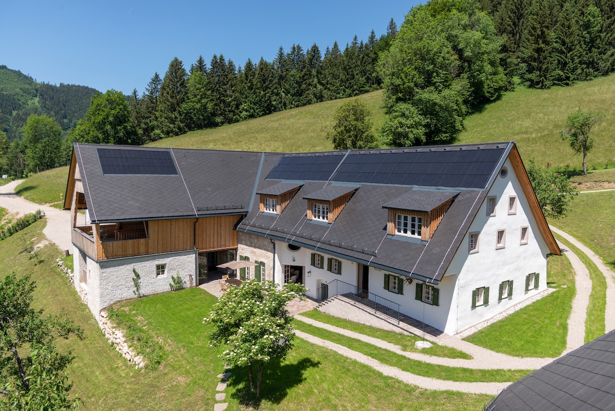 A spacious country house is shown, featuring a mix of modern and traditional architecture. Solar panels are installed on the roof. The surrounding landscape includes lush green hills and trees, enhancing the property's tranquil setting.