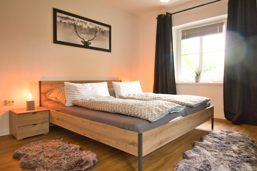 A spacious bedroom is furnished with a large wooden bed and soft bedding. A nightstand with a lamp is placed beside the bed. Natural light enters through a window with minimalistic curtains, and a framed nature photograph adorns the wall, enhancing the room’s peaceful ambiance.
