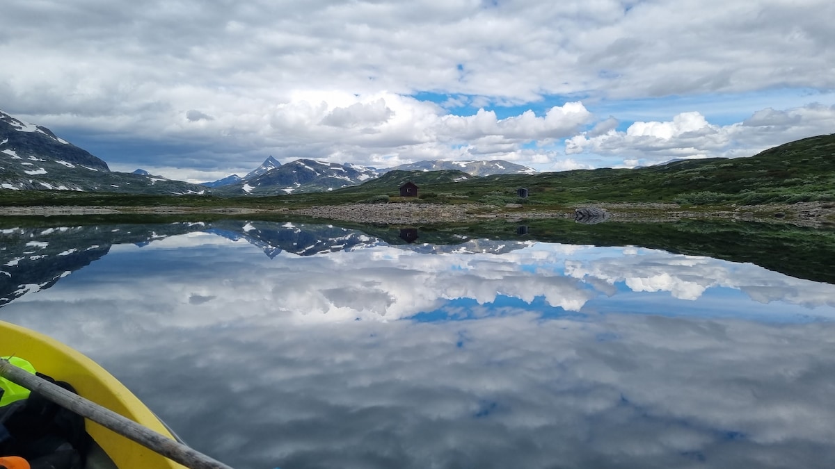 A serene water surface reflects the surrounding mountains and sky, with clouds scattered above. A small cabin is visible on the distant shore, surrounded by lush greenery. The image conveys a sense of tranquility and natural beauty.