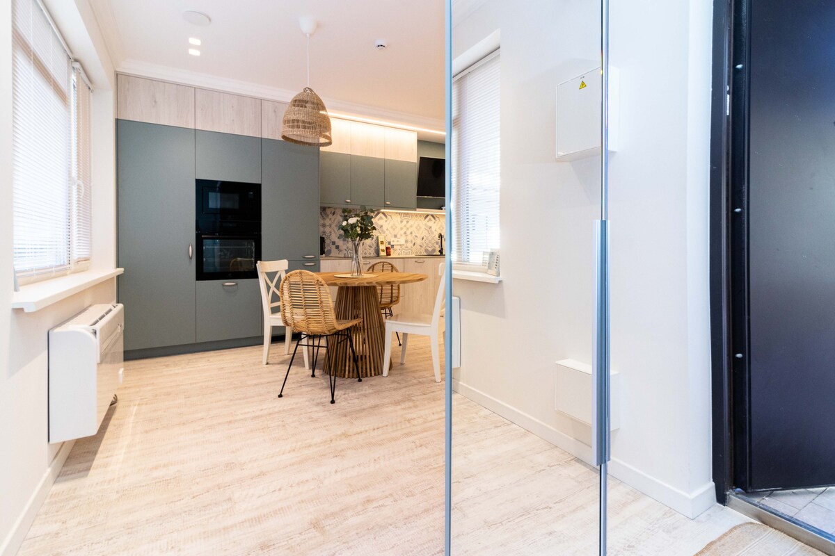 A modern kitchen area is visible, featuring light cabinetry contrasted with dark appliances. A round wooden dining table with four chairs invites casual dining. Natural light floods the space, aided by large windows dressed with light blinds, highlighting clean lines and a neutral color palette.