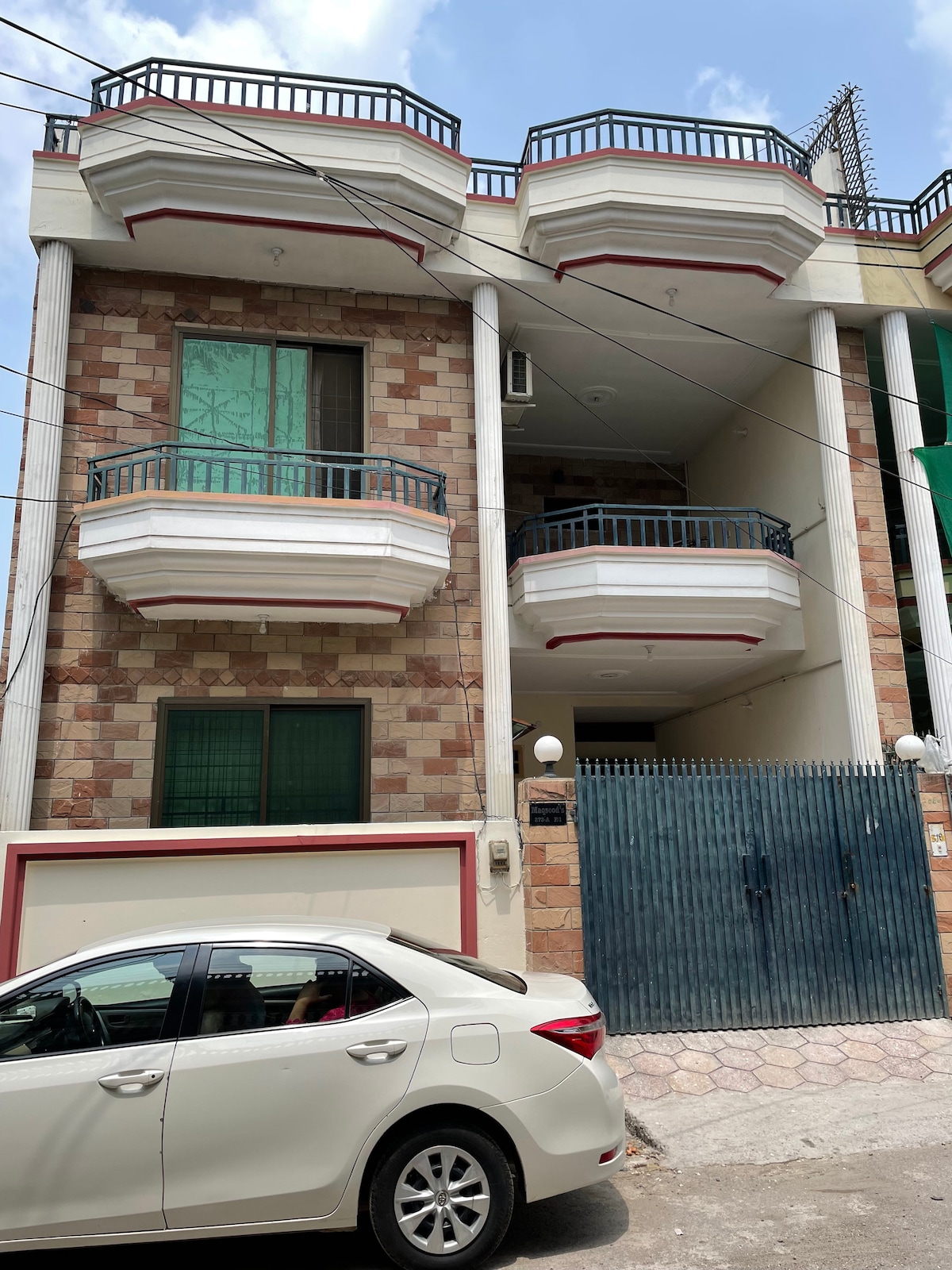 The exterior of the house is showcased, featuring a two-story structure with decorative stone walls. Balconies with railings are visible on both upper levels. A white vehicle is parked in front, while a gated entrance provides access to the property, which is flanked by lush greenery.