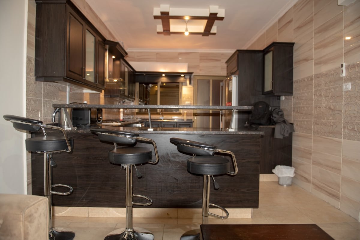 A modern kitchen features dark wooden cabinetry and sleek bar stools positioned at a large island. A stainless steel refrigerator is visible, surrounded by spacious countertops. Neutral tile flooring adds to the contemporary design, enhancing the kitchen's functional layout.