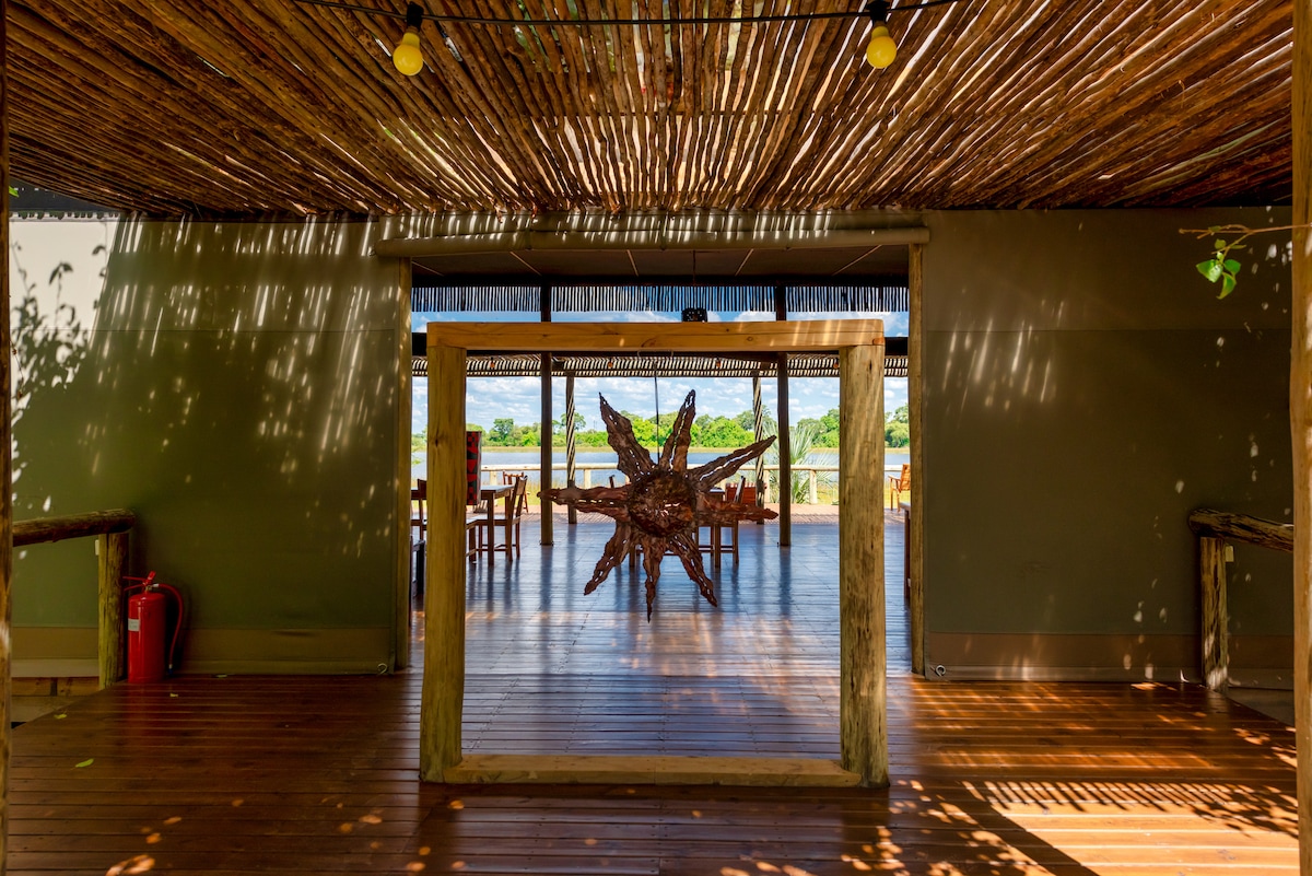 An open space features a wooden floor and a ceiling made of woven materials, allowing light to filter through. A decorative star sculpture is centered in the entryway, with tables and chairs visible in the background, all arranged to face the river view.