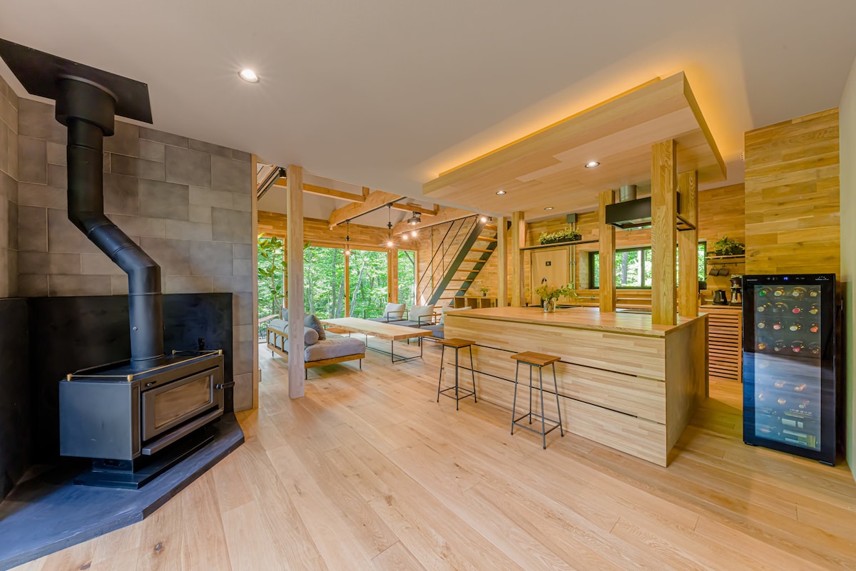 An open living space features natural wood accents and large windows allowing ample natural light. A modern kitchen island with seating faces a cozy wood stove, creating a welcoming gathering area. A sleek wine cooler stands nearby, enhancing the functionality of the space.