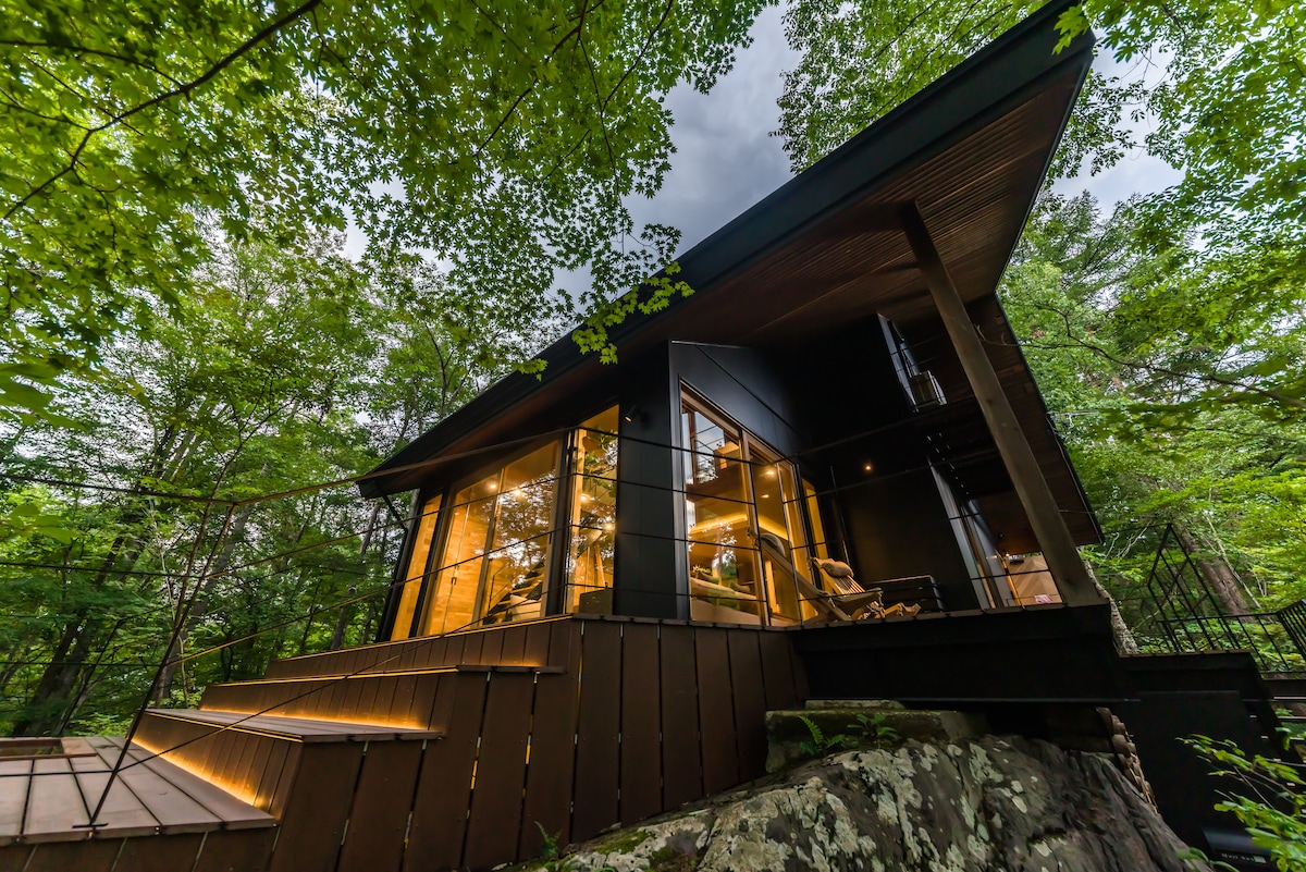 The private villa showcases a modern structure surrounded by lush greenery. Large windows reflect warm indoor lighting, while a wooden deck leads to the entrance. The roof extends outward, enhancing the cabin's unique architectural design within a natural setting.