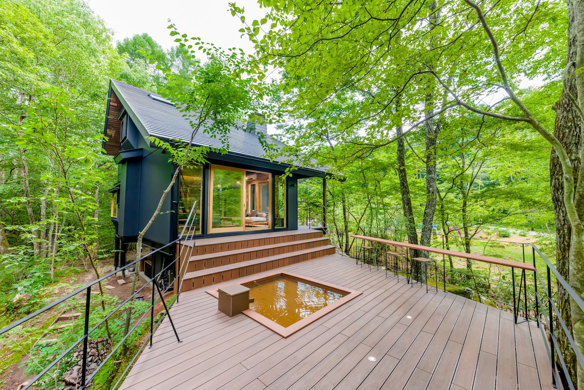 An airy wooden deck extends from the house, surrounded by lush greenery. The structure features large windows, inviting natural light and scenic views of the forest. A small sunken area on the deck offers a space for relaxation.